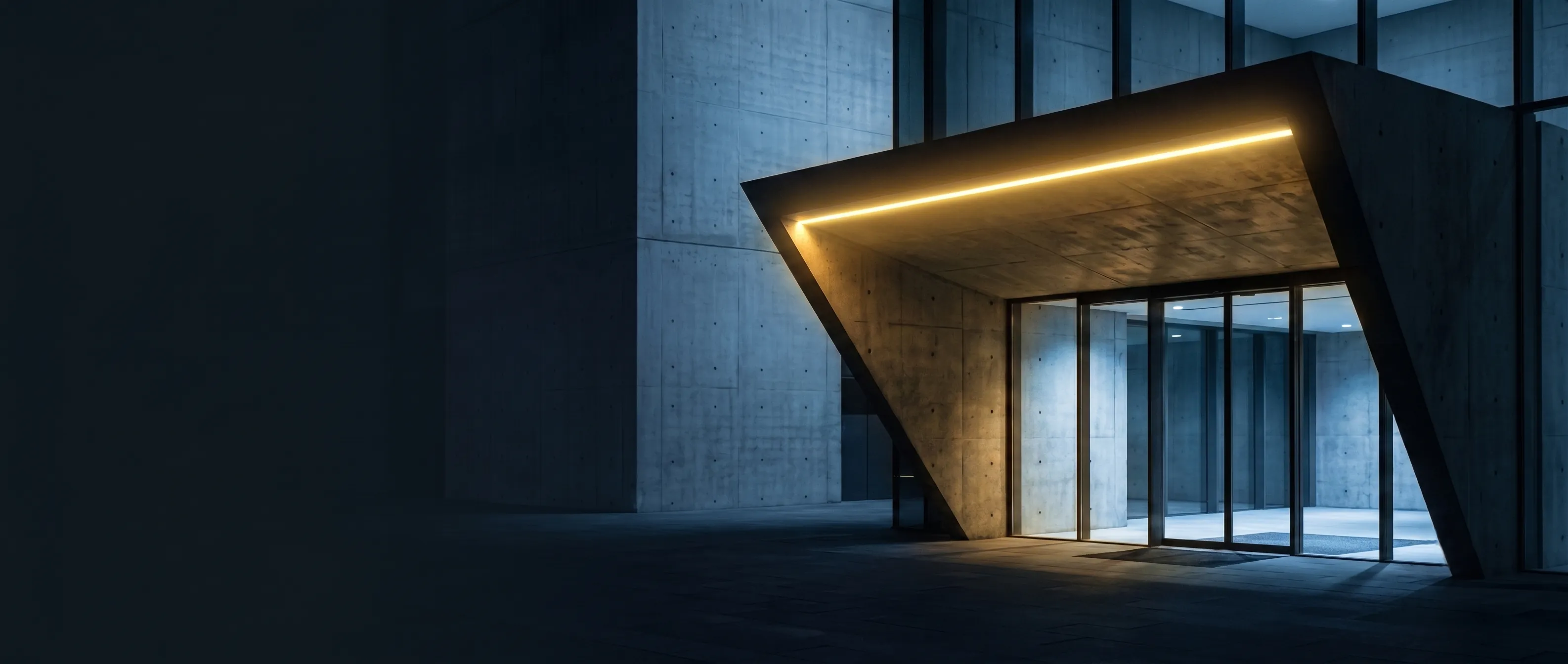Entrée d’un immeuble de béton la nuit : un auvent anguleux en porte-à-faux souligné par une ligne de lumière violette, sous lequel des portes vitrées laissent voir un hall vide éclairé de bleu froid. La moitié gauche reste dans l’ombre.